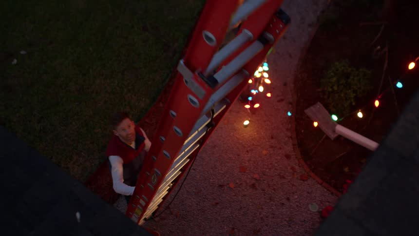 Man operating a ladder outside a house and another person next to him holding lit christmas lights