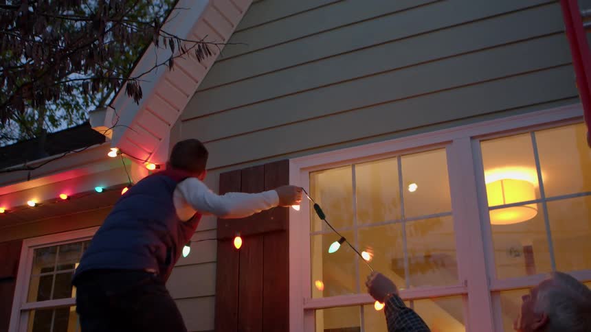 Two people are seen working together to hang colorful christmas lights on the exterior of a house one person stands on a ladder while the other assists from below as they prepare the home for the festive season