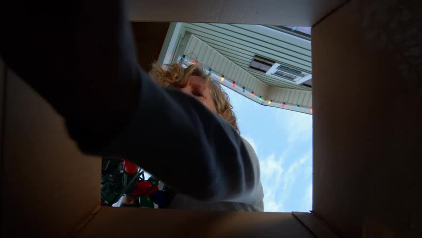 A blonde woman with curly hair looks into an open cardboard box holding a strand of large colorful christmas lights with a bright blue sky and part of a house visible above her