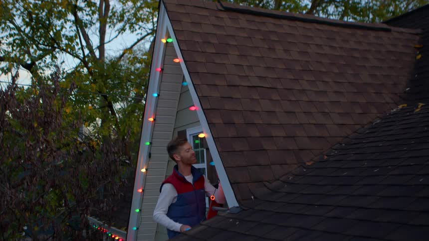 A man stands by the roof of an aframe house hanging colorful string lights as evening sets in