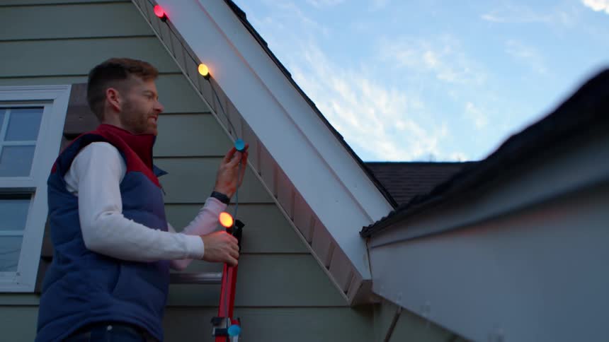 A man standing on a ladder diligently attaching colorful string lights to the eaves of a house the bright blue sky in the background suggests it is daytime and he appears focused on his task