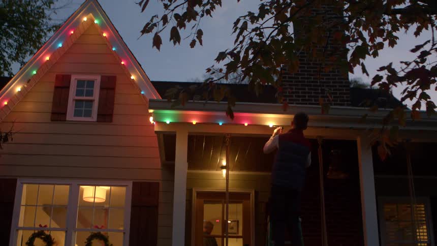 A man stands on a ladder hanging colorful lights from the roof of a house with a lit wreath visible in a window below