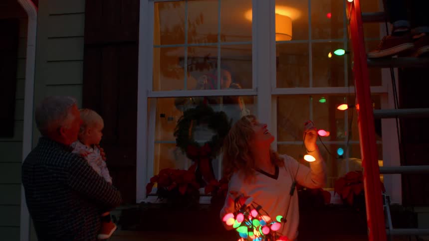 Man on a ladder decorating his house with christmas lights accompanied by other people at dusk