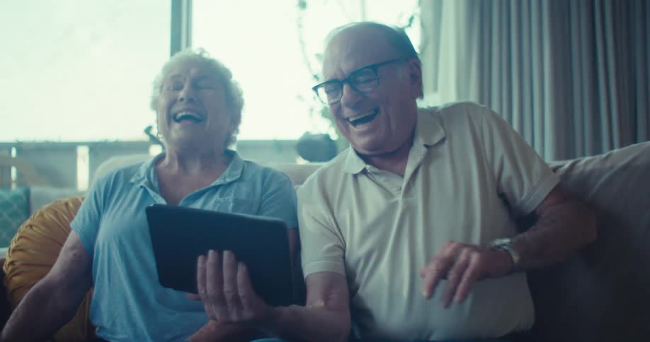 Elderly couple sitting on the couch and laughing while interacting with the tablet