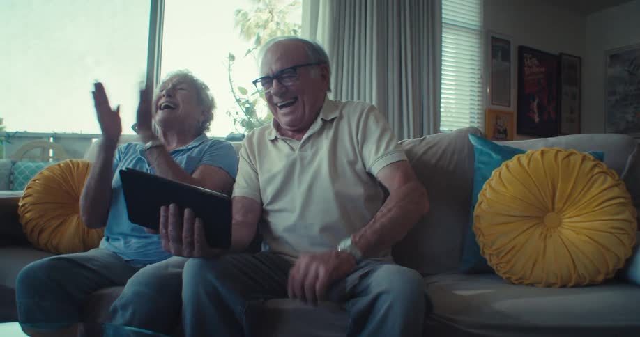 An elderly couple laugh joyfully while looking at a tablet on the couch