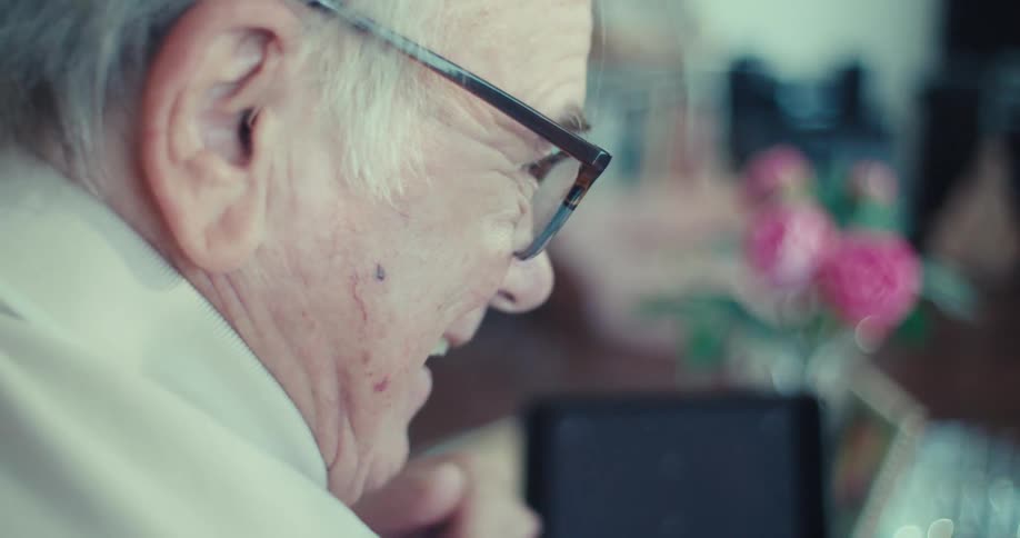 An elderly man holding a black tablet with six white tracking markers on its dark screen speaks to an elderly woman in a cozy home setting