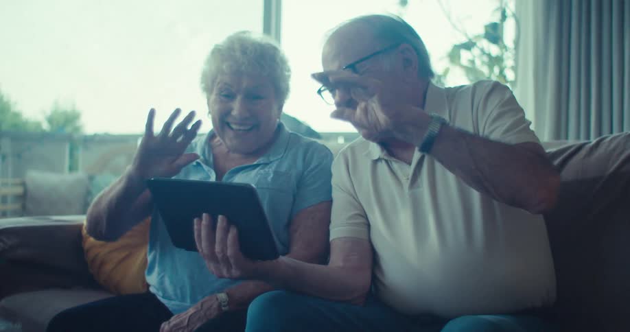 An elderly couple waves at a tablet while video chatting from their living room