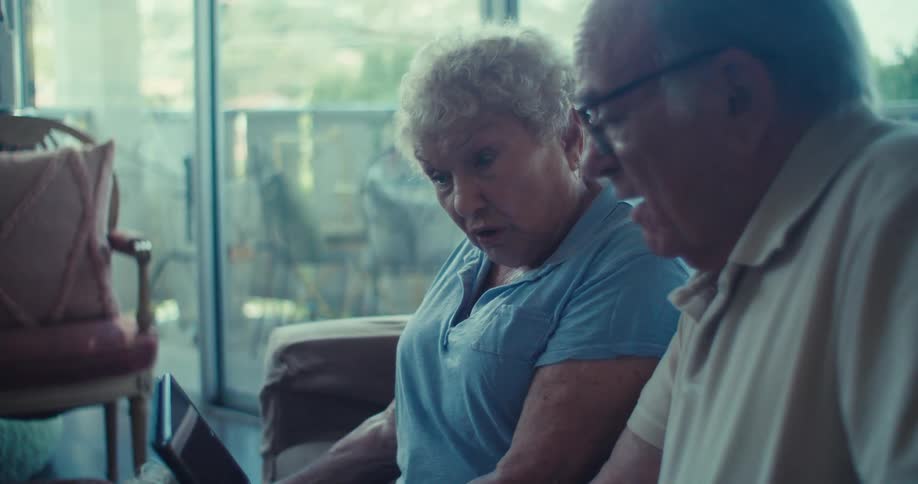 An elderly couple sits indoors focused on something on a tablet