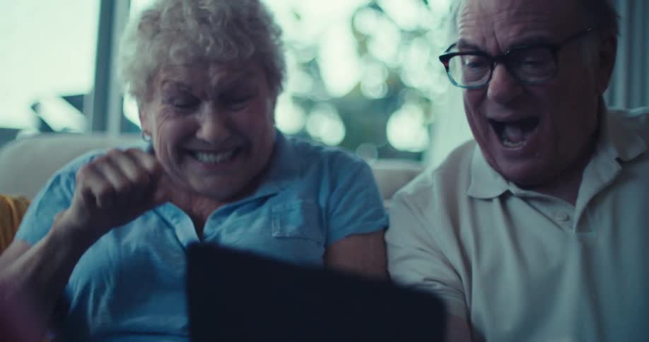 Elderly couple with happy expression and interacting with tablet while sitting on sofa in living room