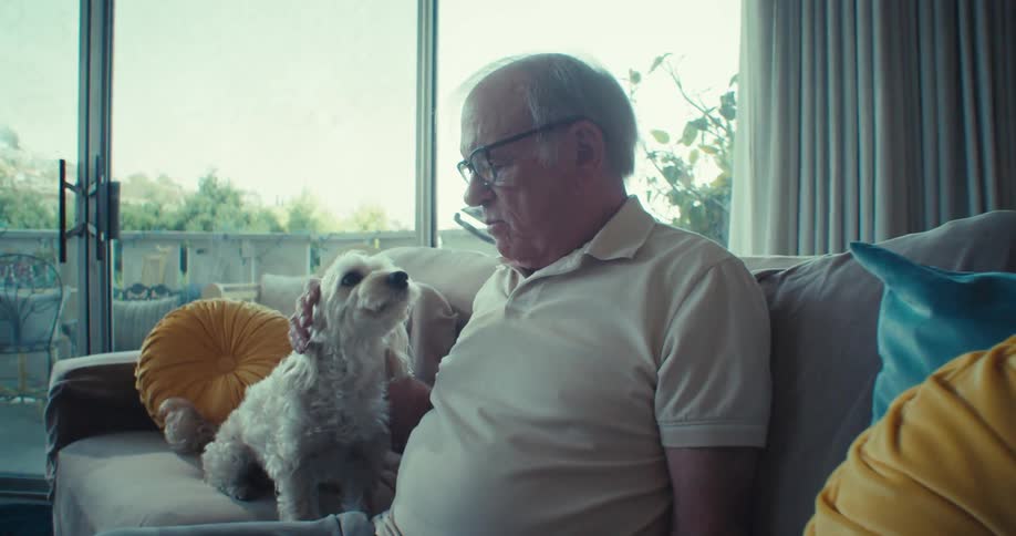 Elderly man sitting on the sofa in the living room and interacting with a white dog next to him