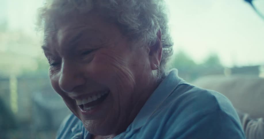 An elderly couple sits side by side in a brightly lit room laughing warmly as they face forward