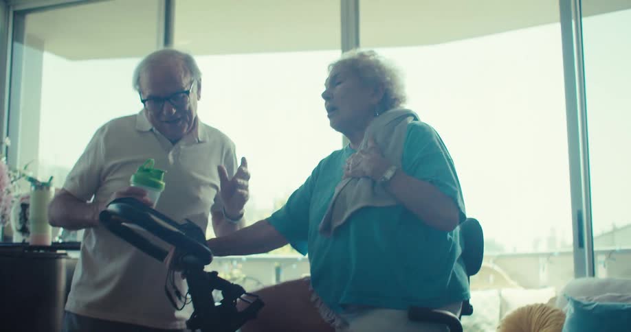 Elderly woman exercises on a stationary bike while man supports her with a drink in a cozy living room