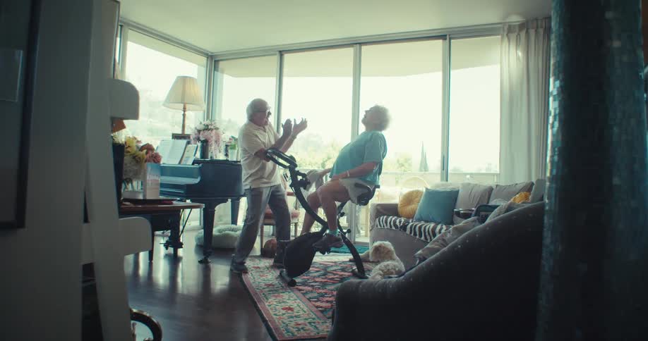 Elderly man encouraging his elderly wife by handing her a bottle of water while doing exercise bike in the living room