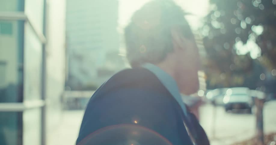 A greyhaired man in a suit jacket and collared shirt holds a black cellphone to his ear reacting with joyful surprise on a sunlit city street