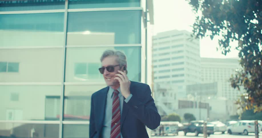 A stylish greyhaired man in a suit and sunglasses confidently walks down a city street while engrossed in a phone conversation the bright sunny day highlights the modern architecture and bustling urban environment around him
