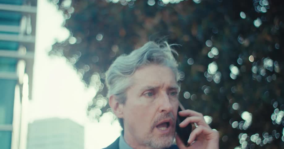 An older businessman in a suit and tie walking outdoors and talking excitedly on his phone he appears to be reacting positively to the conversation