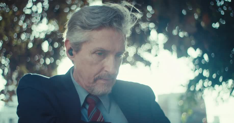 An older businessman wearing a suit and tie attempting to put an earbud into his ear he struggles slightly with the small device looking focused and a bit contemplative amidst what appears to be an outdoor sunny setting
