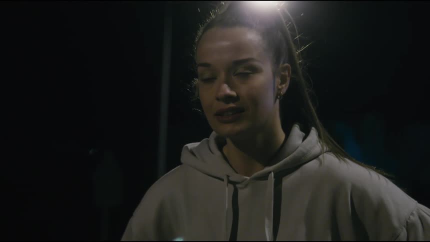 A woman with long dark hair pulled back wearing a lightcolored hoodie she appears to be outside at night with a bright light source possibly a street lamp visible in the background creating a strong backlight effect