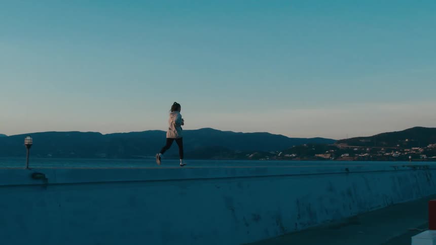 A woman with long braided hair runs on a concrete platform separating the shoreline with a series of boats on the right side as the sun rises