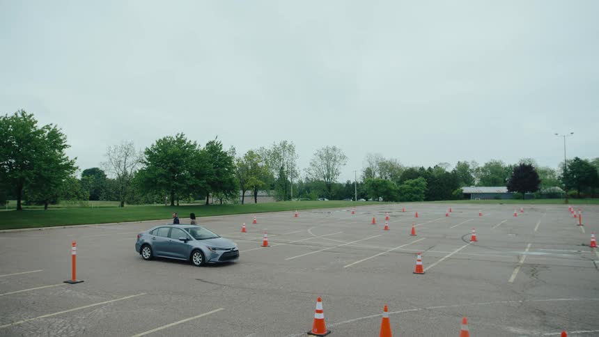 Two mean stand next to a car in a large parking lot set up with orange cones for a driving test on an overcast day