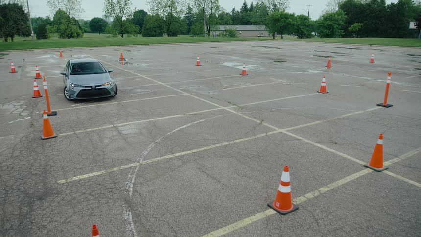 A car drives through an empty parking lot where a series of cones are placed at midday