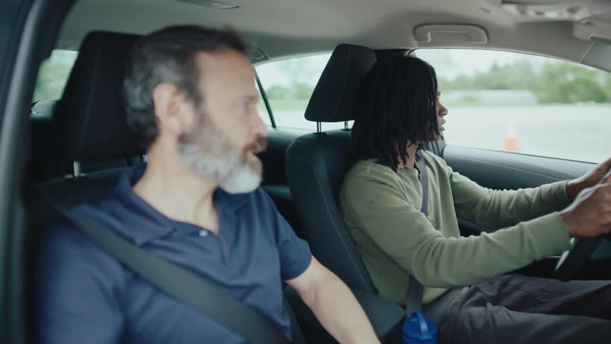 A young black man practices driving with a male instructor in a vehicle during daytime