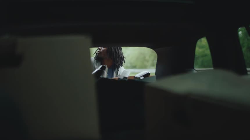 A dark car looking out through the rear window shows a person with long dark hair possibly dreadlocks and a lightcolored top placing items into the back of the vehicle they look up briefly towards the camera with a serious expression