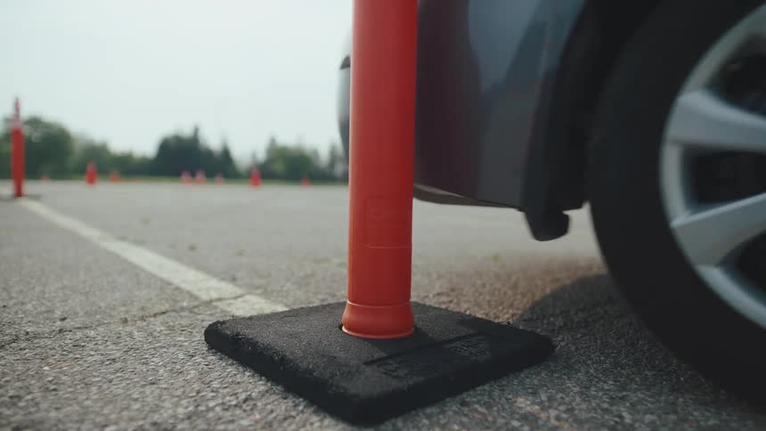 On a driving course a cars front wheel runs over the base of an orange pylon pushing it forward