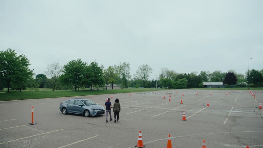 A gray car positioned in a large empty parking lot marked with orange cones suggesting a driving practice area two individuals then walk into the frame and approach the vehicle