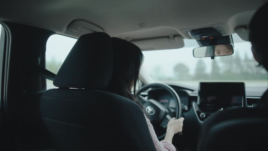A young asian woman steers while driving with a male driving instructor at practice during daytime