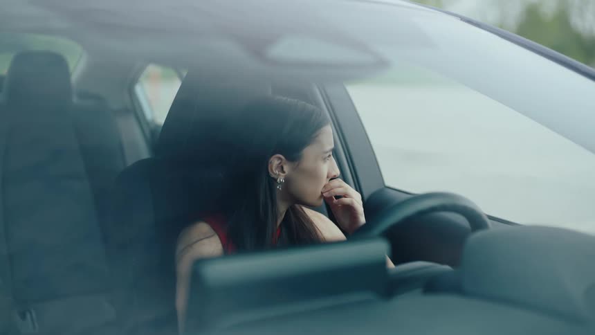 A young woman sitting in the drivers seat of a car viewed through the windshield she appears to be deep in thought or possibly distressed as she repeatedly touches her chin and face her expression suggests contemplation or worry