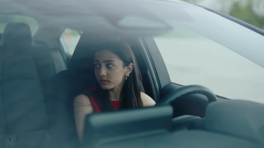 A woman with long dark hair dressed in a red sleeveless top sitting in the drivers seat of a car she appears to be concentrating while looking straight ahead with a serious expression