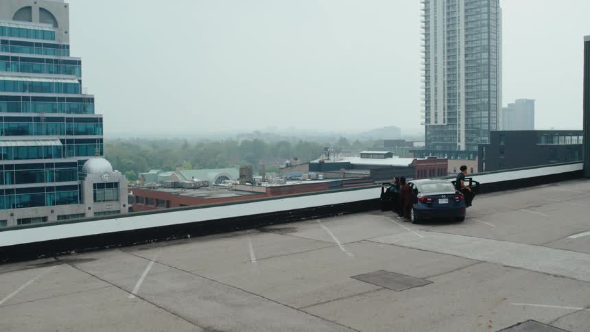 A black car pulling into a parking spot on a rooftop in a city several people then exit the car and gather around it looking out at the urban landscape