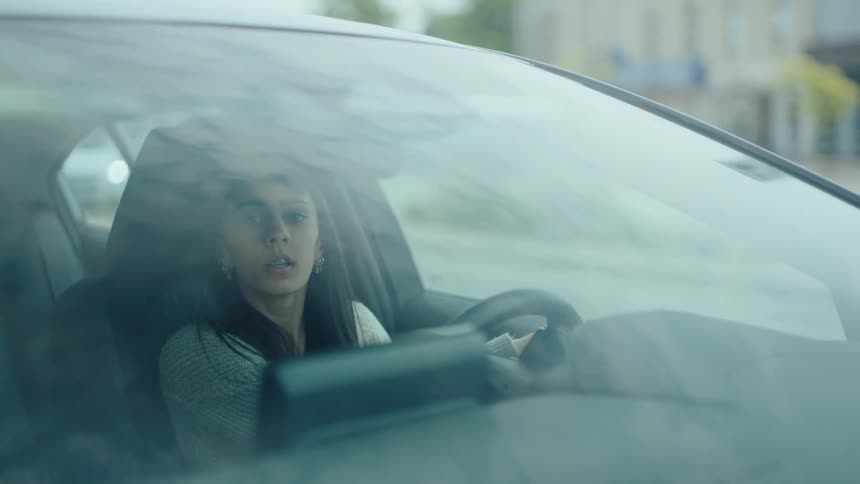 A young woman sitting in the drivers seat of a car appearing focused and a bit anxious as she looks around her facial expressions suggest concentration possibly while learning to drive or navigating a challenging situation