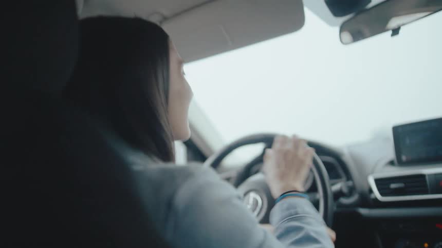 A person with a long ponytail wearing a light blue shirt driving a car from a drivers perspective they turn their head to the right looking into the passenger seat as if talking to someone