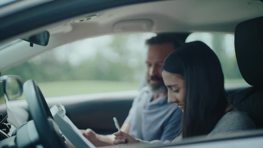 A male driving instructor congratulates a female driver after driving practice in a vehicle during daytime
