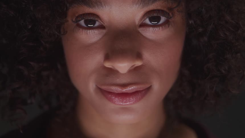 A young woman with long black curly hair wearing lipstick and soft makeup smiles warmly