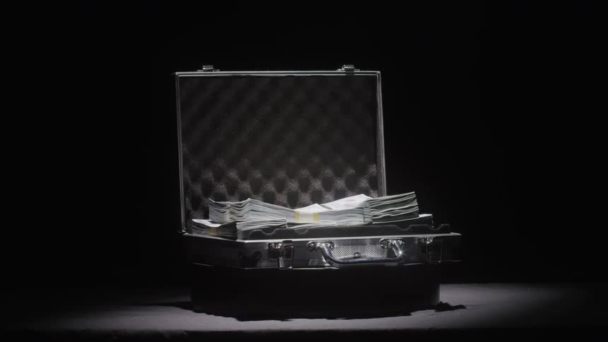A metal briefcase full of neatly stacked cash sits open in a spotlight