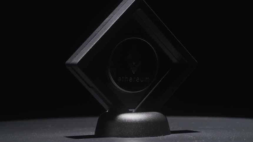 A stylized dark transparent display stand featuring the ethereum logo and text slowly rotates in a spotlight against a black background the objects subtle reflections highlight its angular design presenting it as a futuristic