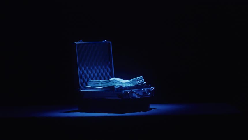 An open suitcase filled with money sits in a spotlight against a dark background