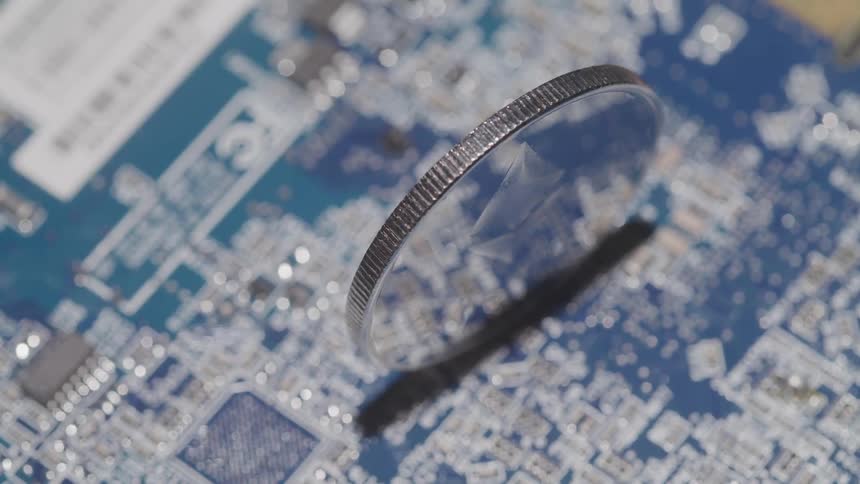 A shiny and reflective coin is balanced on its edge atop a printed circuit board