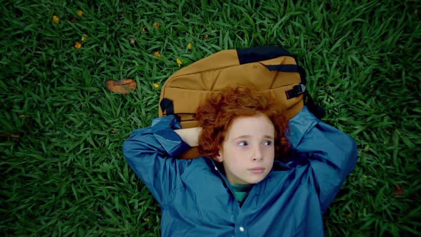 A child with red curly hair lies on a patch of lush green grass their head resting on a brown backpack they look directly upwards their face conveying a sense of thoughtful contemplation