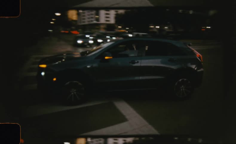 A dark suv drives through a city intersection at night captured in a grainy filmstyle shot from a moving perspective
