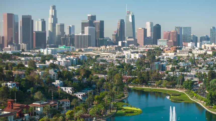 An aerial timelapse captures the bustling downtown los angeles skyline as seen over a serene city park and lake on a sunny day