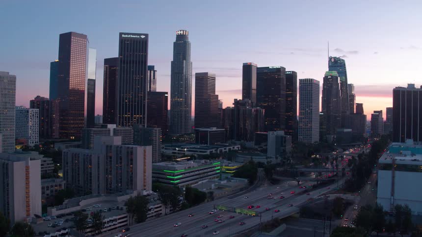 Downtown los angeles glows at dusk as traffic streams along the freeway beneath a skyline of glass towers and cranes