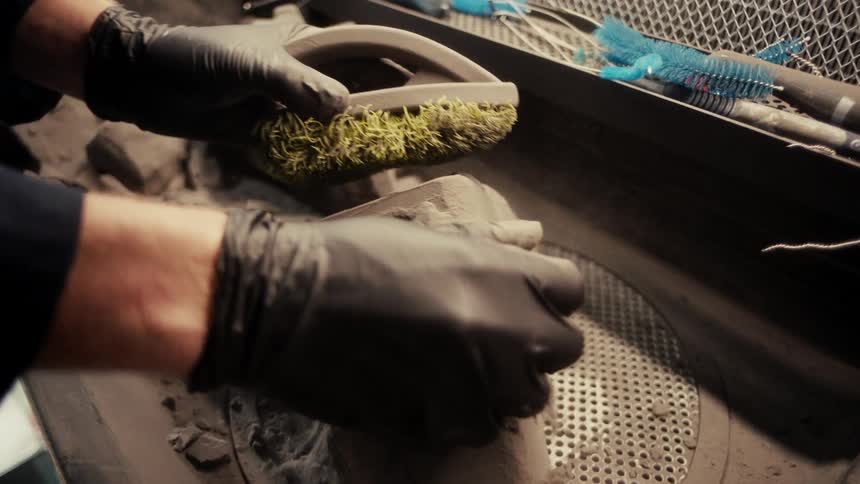 A person wearing black gloves brushes dirt off an object in a dimly lit workshop