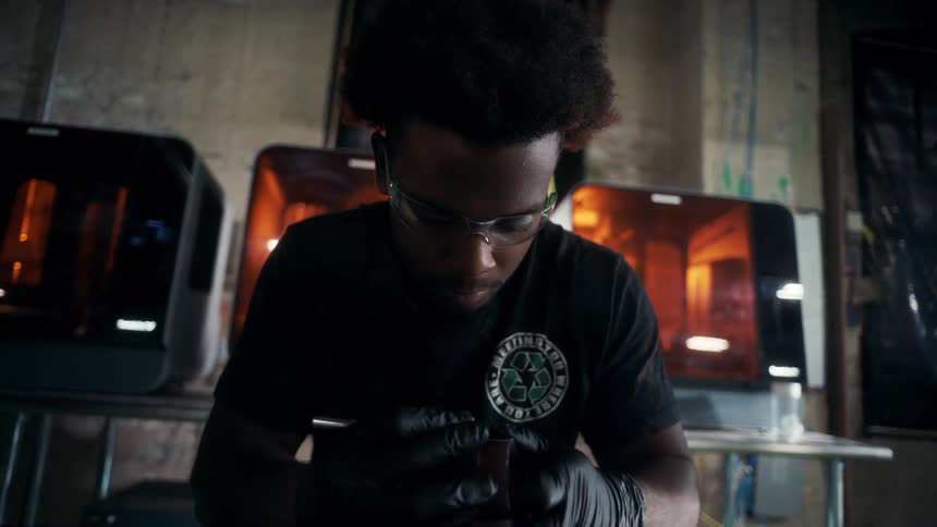 A person wearing black gloves works on a small component in a workshop filled with tools cables and equipment