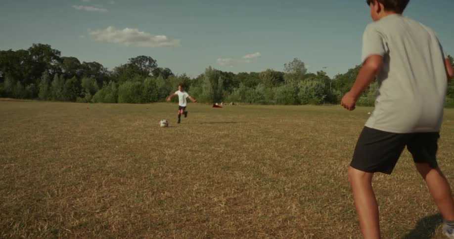 Two boys practice soccer in a large dry field with one kicking the ball towards the other who then controls it with his knee