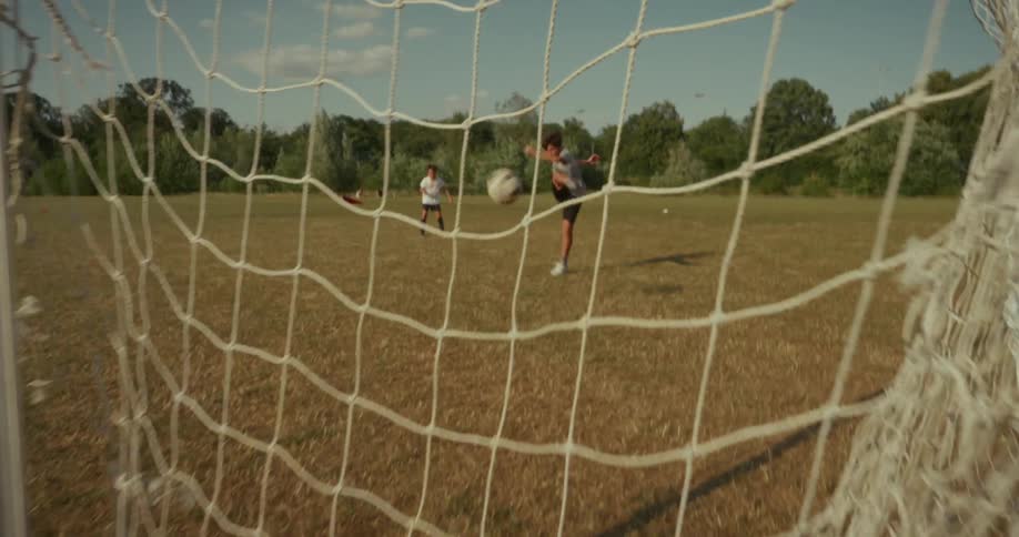 A wide shot through a soccer goal shows two players on a dry grassy field with one player dribbling the ball the player in the foreground kicks the ball and it rolls offscreen towards the right