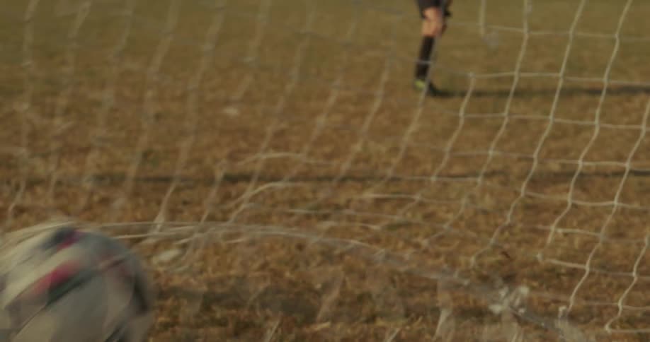 A boy is kicking a ball into a soccer net outdoors
